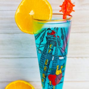 A blue AMF cocktail drink garnished with an orange slice that's in a vintage pirates motif glass