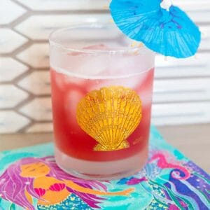 Bay Breeze Cocktail in a glass emblazoned with a gold shell and garnished with a small umbrella on a mermaid napkin