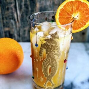 Vertical photo of a Brass Monkey Cocktail in a Mardis Gras style tumbler garnished with an orange. full orange in the background.