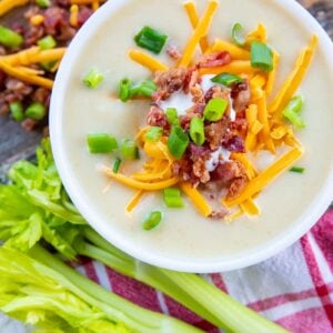 close up Super thick Cream of Celery Soup topped with shredded cheese, bacon bits and green onions in a white soup bowl. Raw celery stem and some toppings on its background