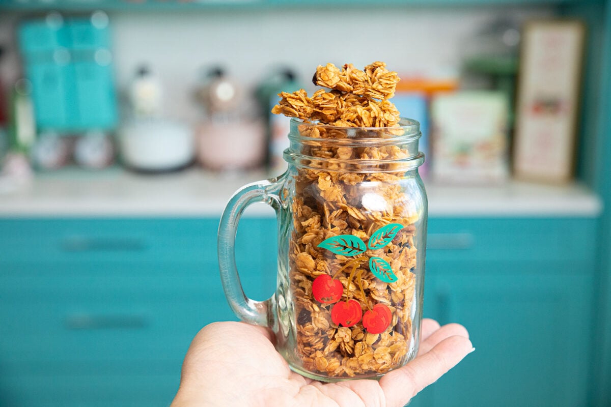 Granola Clusters in a mason jar mug being held up on a flat palm. 