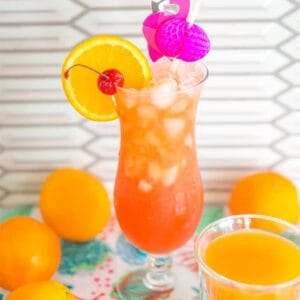 Hurricane Cocktail in a hurricane glass on a counter top surrounded by oranges with a side of passion fruit juice.