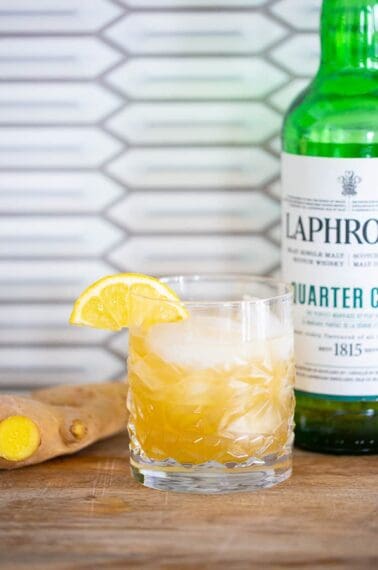 a glass of penicillin cocktail garnished with a lemon slice, a piece of ginger and a bottle of laphroaig