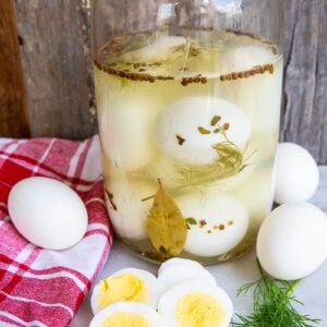 close up Pickled Eggs in sterilized 2-litre glass jar. Red checkered table cloth, boiled eggs and sprigs fresh dill around the jar