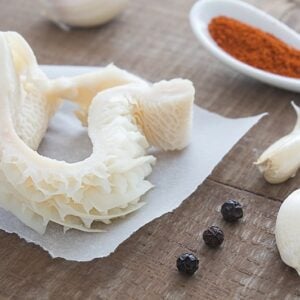 close up raw tripe on a parchment paper, black pepper corns, garlic cloves and powder seasoning on spoon beside it