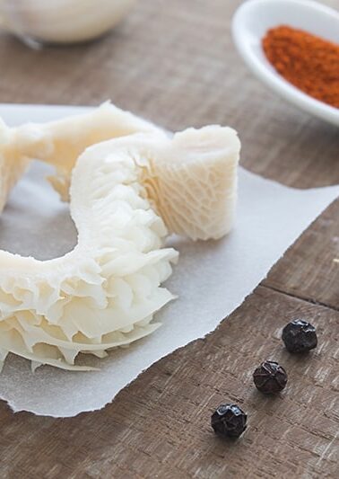 close up raw tripe on a parchment paper, black pepper corns, garlic cloves and powder seasoning on spoon beside it