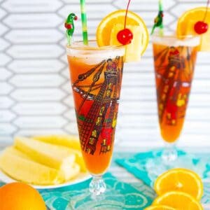 Rum Runner Cocktail in a tall pirate designed pilsner glass on a turquoise tablecloth surrounded by oranges, pineapple wedges and banana slices.