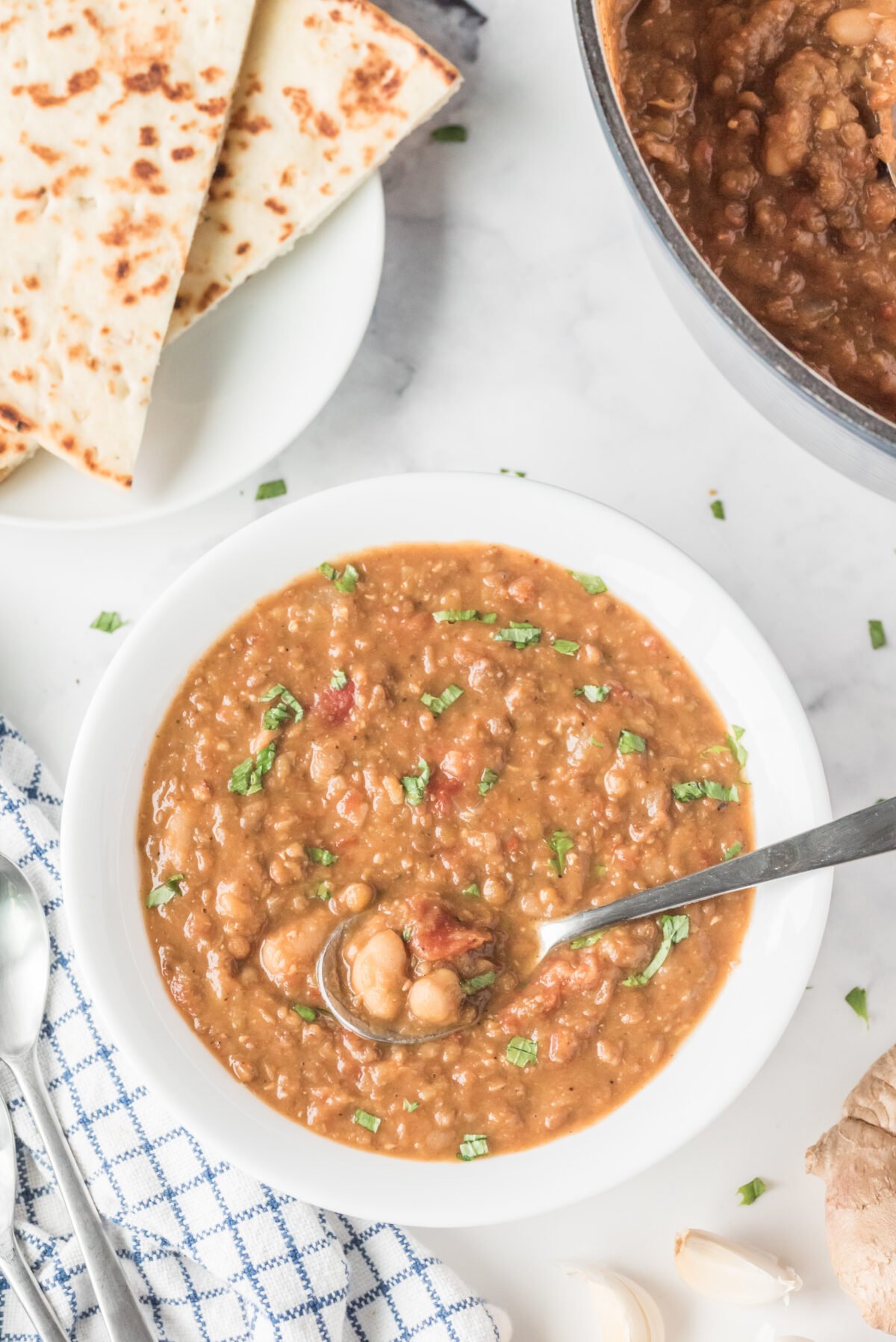 spicy bean and lentil soup in a white bowl with a spoon with some naan bread on a plate beside the bowl