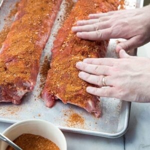 Raw pork ribs being prepared with rib rub on a baking sheet